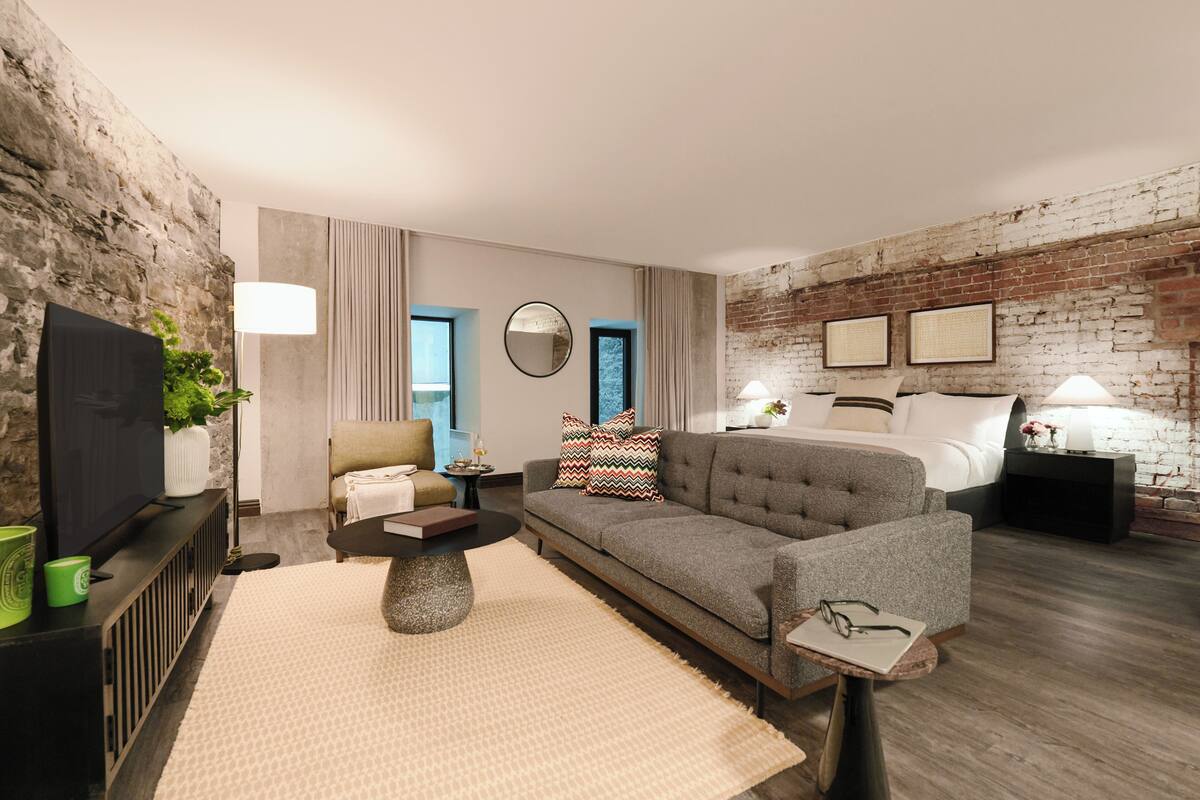 A modern hotel room with a stone wall, a grey sofa, a bed with white linens, a television, and a small table with a lamp.