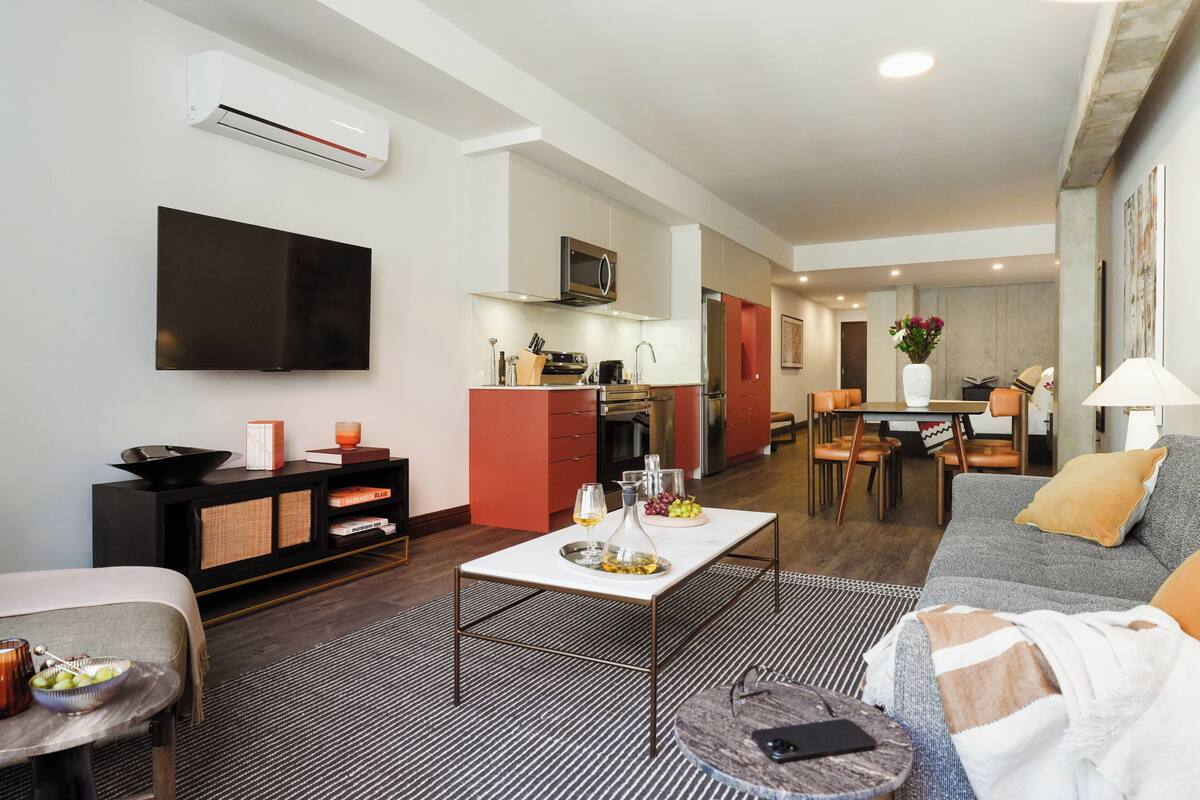 A modern living room with a flat-screen TV, a dining area with a table and chairs, a kitchen with a microwave and oven, and a sofa with a throw blanket.