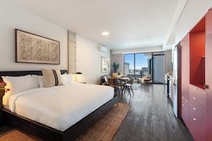 A modern hotel room with a large bed, a dining area, and a view of the city.