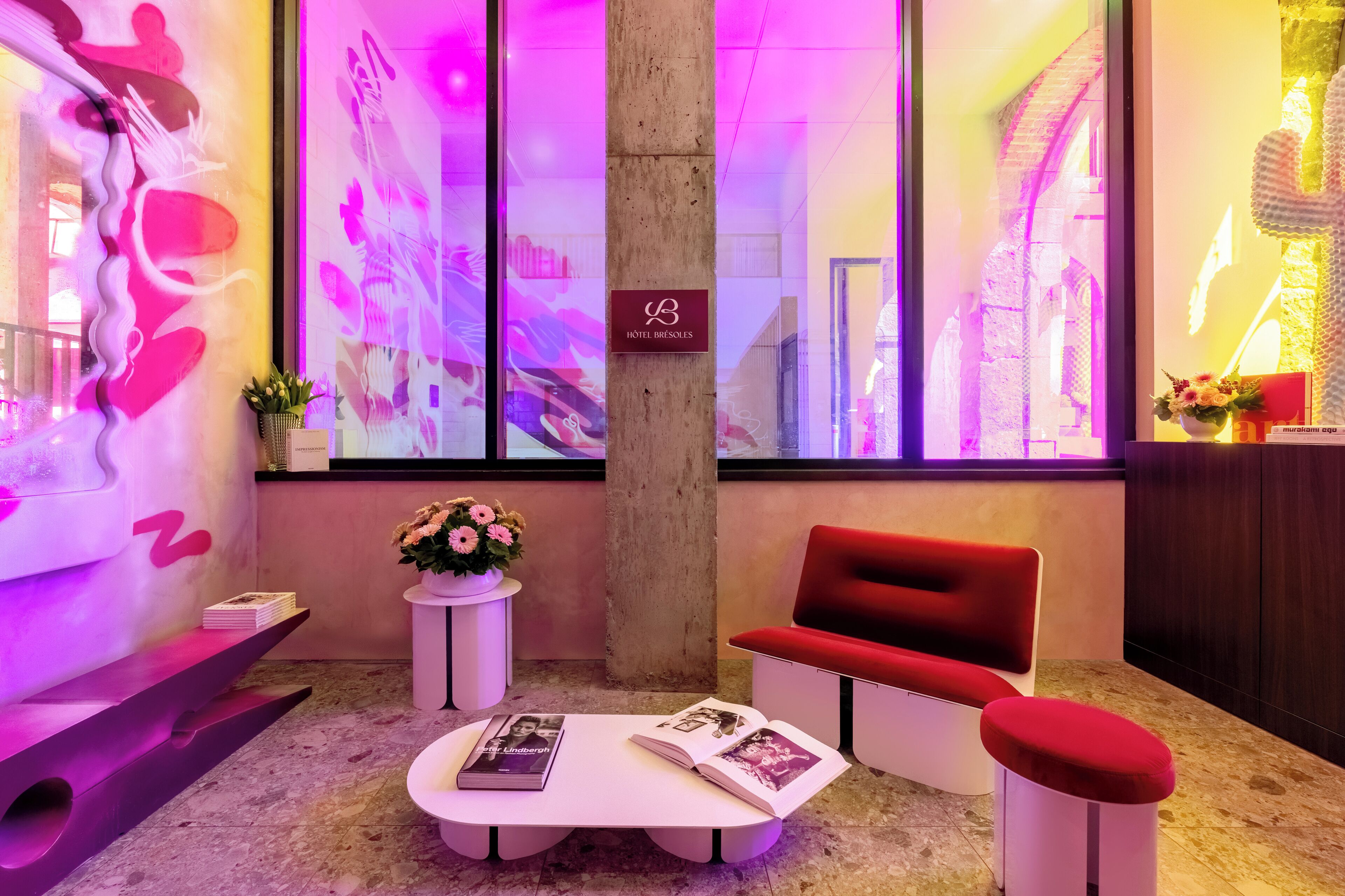 Un hall d’entrée moderne avec un canapé rouge, une table basse blanche sur laquelle sont posés des magazines, une petite table d’appoint avec une plante et une grande fenêtre ornée d’œuvres d’art abstraites.