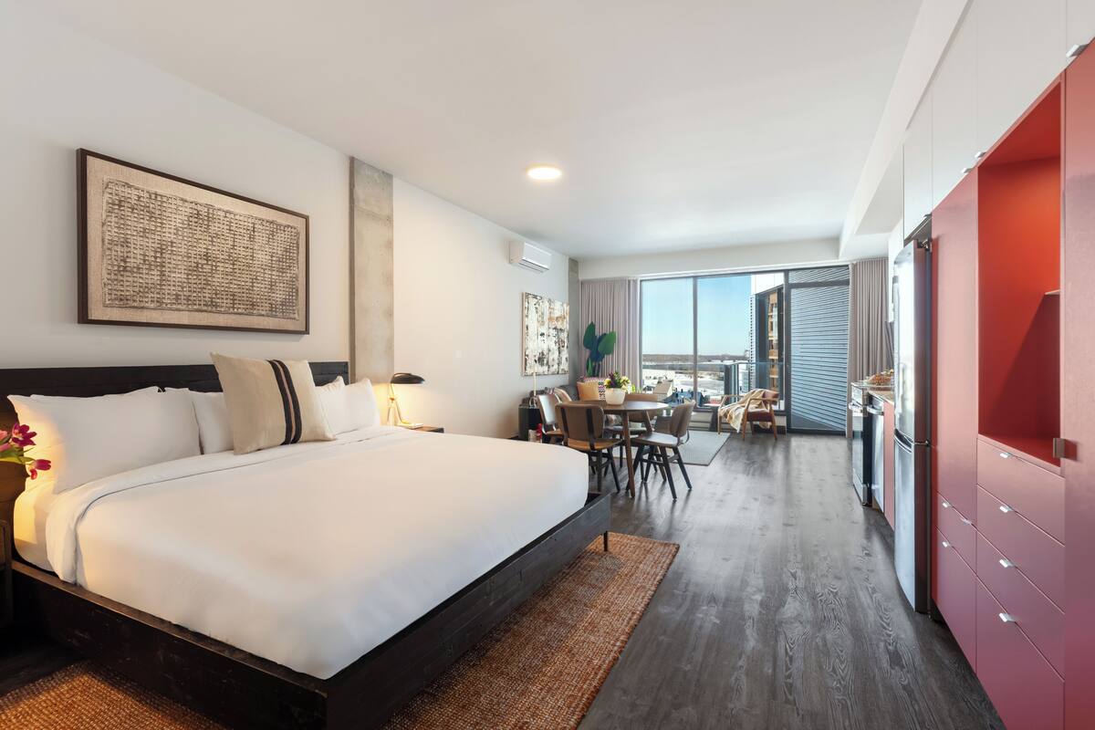 A modern hotel room with a large bed, a dining area, and a view of the city.