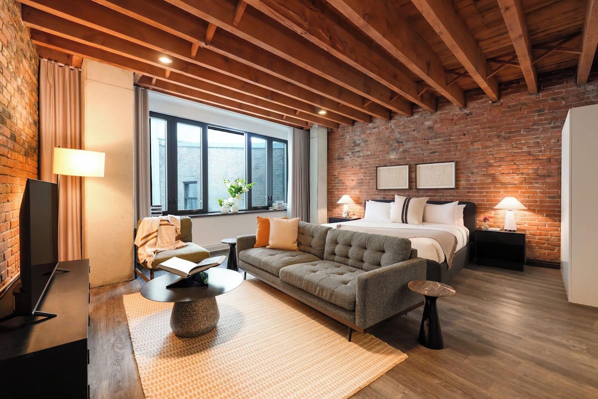 A modern bedroom with a brick wall, a large window, a bed, a sofa, and a TV.