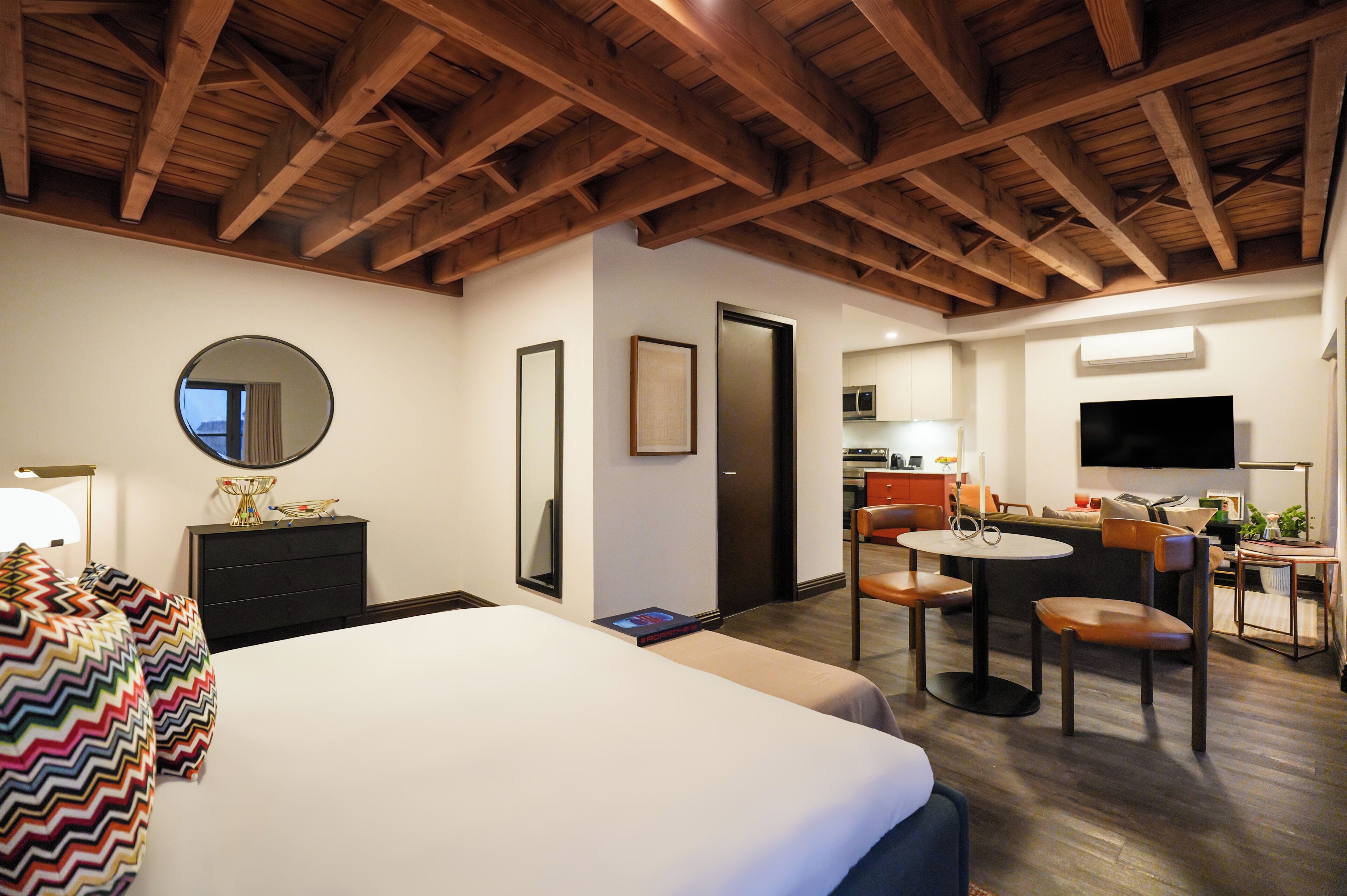 Una habitación de hotel con cama, zona de comedor con sillas, televisor y vigas de madera en el techo.