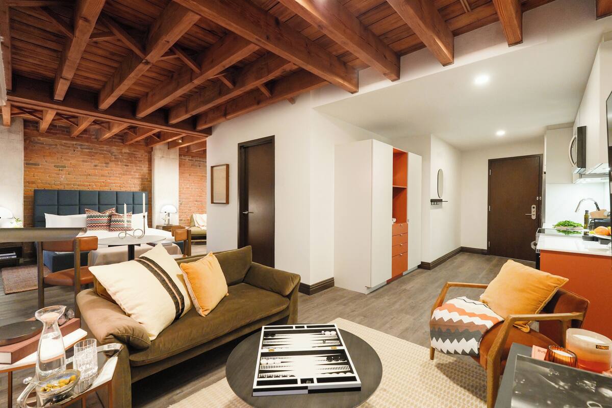 A modern living room with a brick wall, wooden ceiling beams, a sofa, a dining table, and a kitchen area.