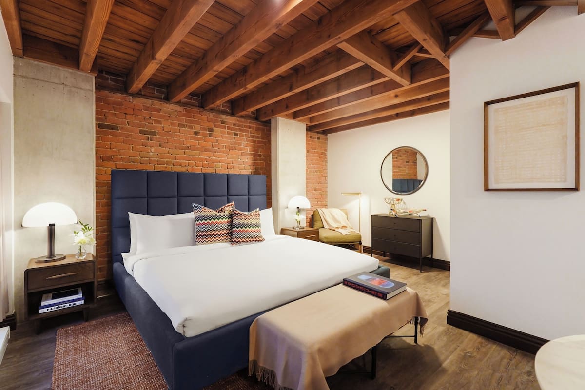 A bedroom with a large bed, wooden beams, a brick wall, a round mirror, and a framed artwork on the wall.