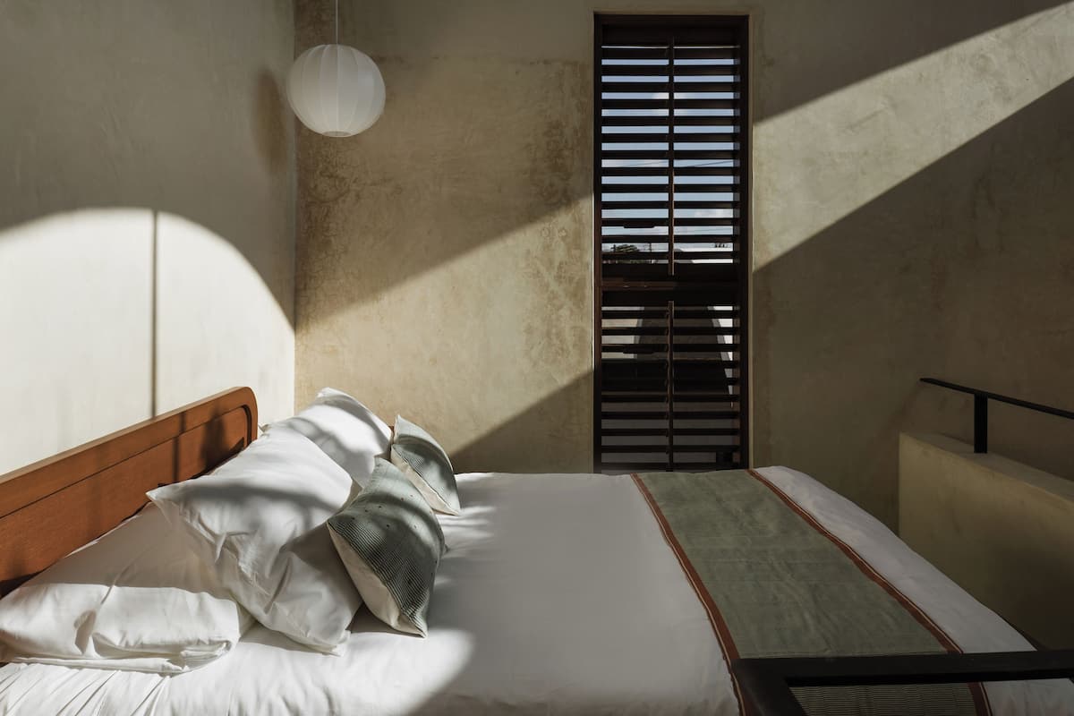 Ein Zimmer mit einem Bett, einem Fenster mit Jalousien, einer Pendelleuchte und einem Teil eines Wandbildes.
