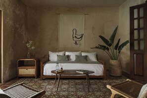 A room with a bed, a small table, a chair, a large plant, and a wall tapestry. - Serena Hotelito Boutique (Mérida)