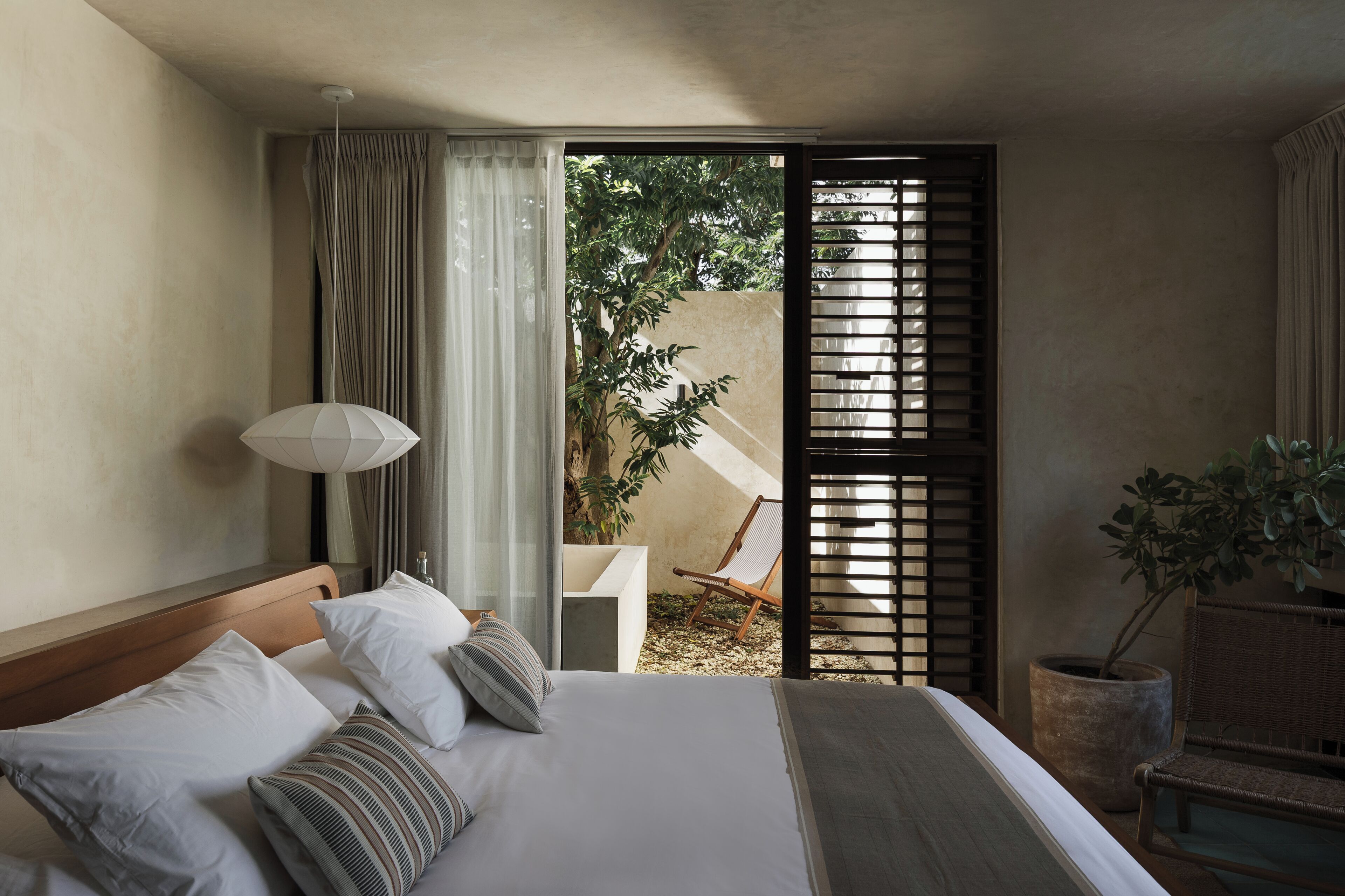 A bedroom with a bed, a nightstand, a lamp, a window with curtains, and a view of the outdoors.
