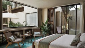 Een slaapkamer met een groot bed, een zithoek met stoelen, een kleine tafel en een groot raam met uitzicht op een tuin.