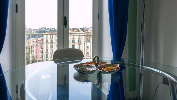 A room with a glass table, a chair, and a view of buildings through a window with blue curtains. - Elegant Apartment for 4 in Chiaia - Capri View Terrace (Napoli)