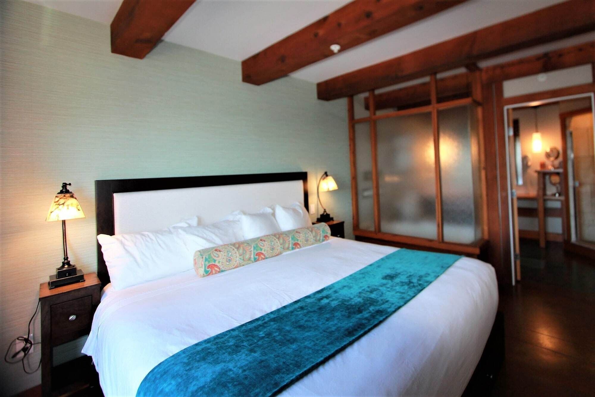 A hotel room with a bed, two bedside lamps, a wooden ceiling, and a sliding door leading to a bathroom.
