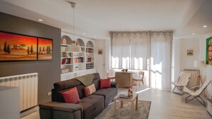 Ein Wohnzimmer mit einer Couch, einem Couchtisch und Sesseln, einem Bücherregal sowie einem Essbereich mit Tisch und Stühlen.