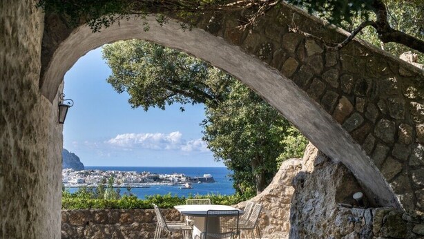 An outdoor patio with a stone archway, a table, and chairs, overlooking a coastal town and the sea. - Villa Grotta Leone (Campania)