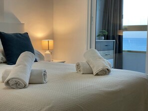 Ein ordentlich bezogenes Bett mit aufgerollten Handtüchern, einer Nachttischlampe und Blick auf das Meer durch ein Fenster.