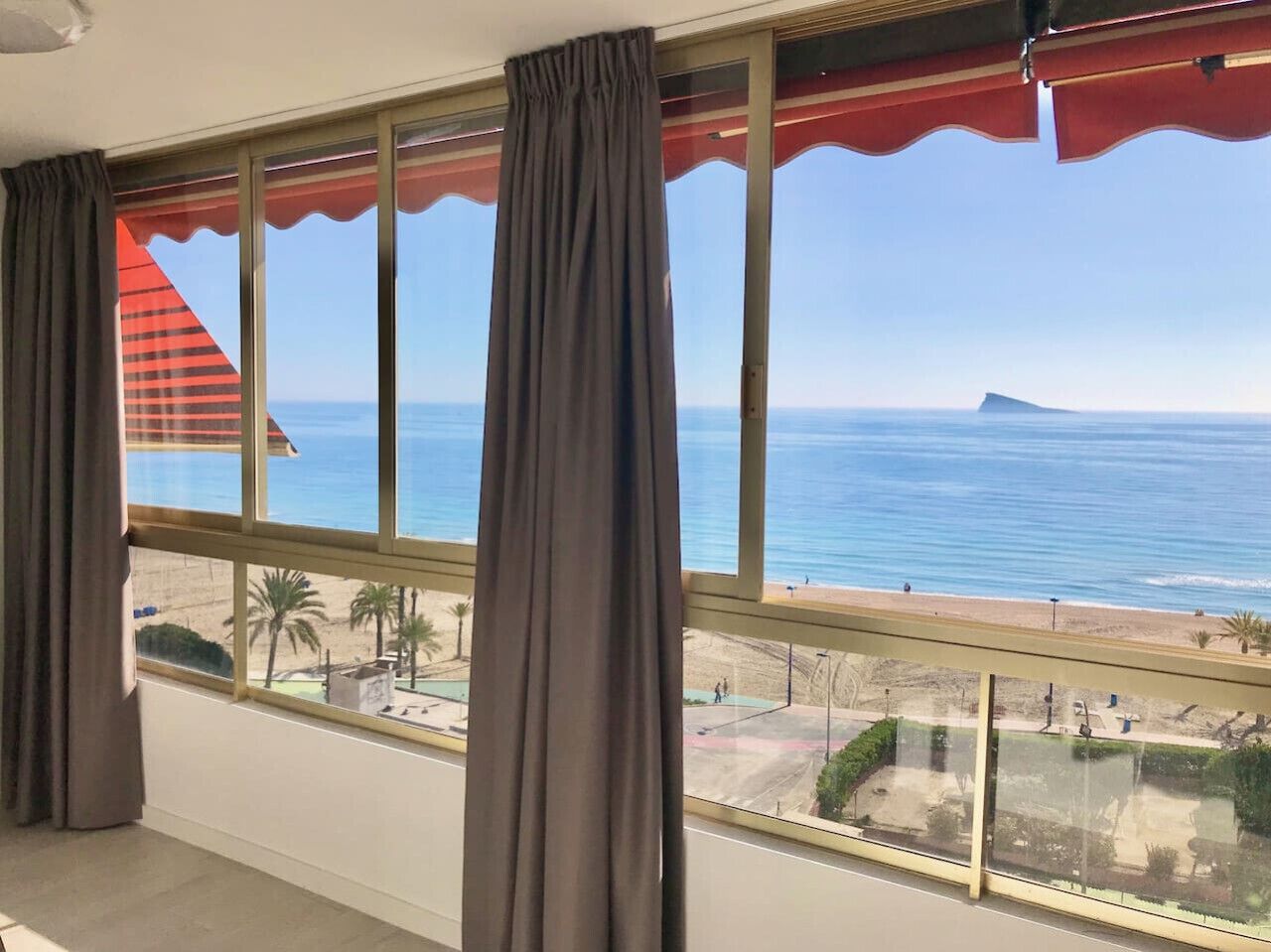 A room with large windows overlooking a beach, a balcony with a railing, and a view of the sea.