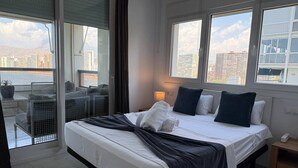 A hotel room with a bed, a nightstand, a lamp, and a view of the city through large windows.