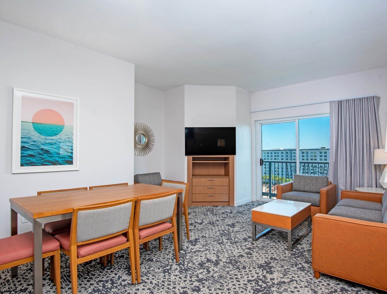 Habitación de hotel con mesa de comedor, sofá, televisor y un amplio ventanal con vista a la ciudad.