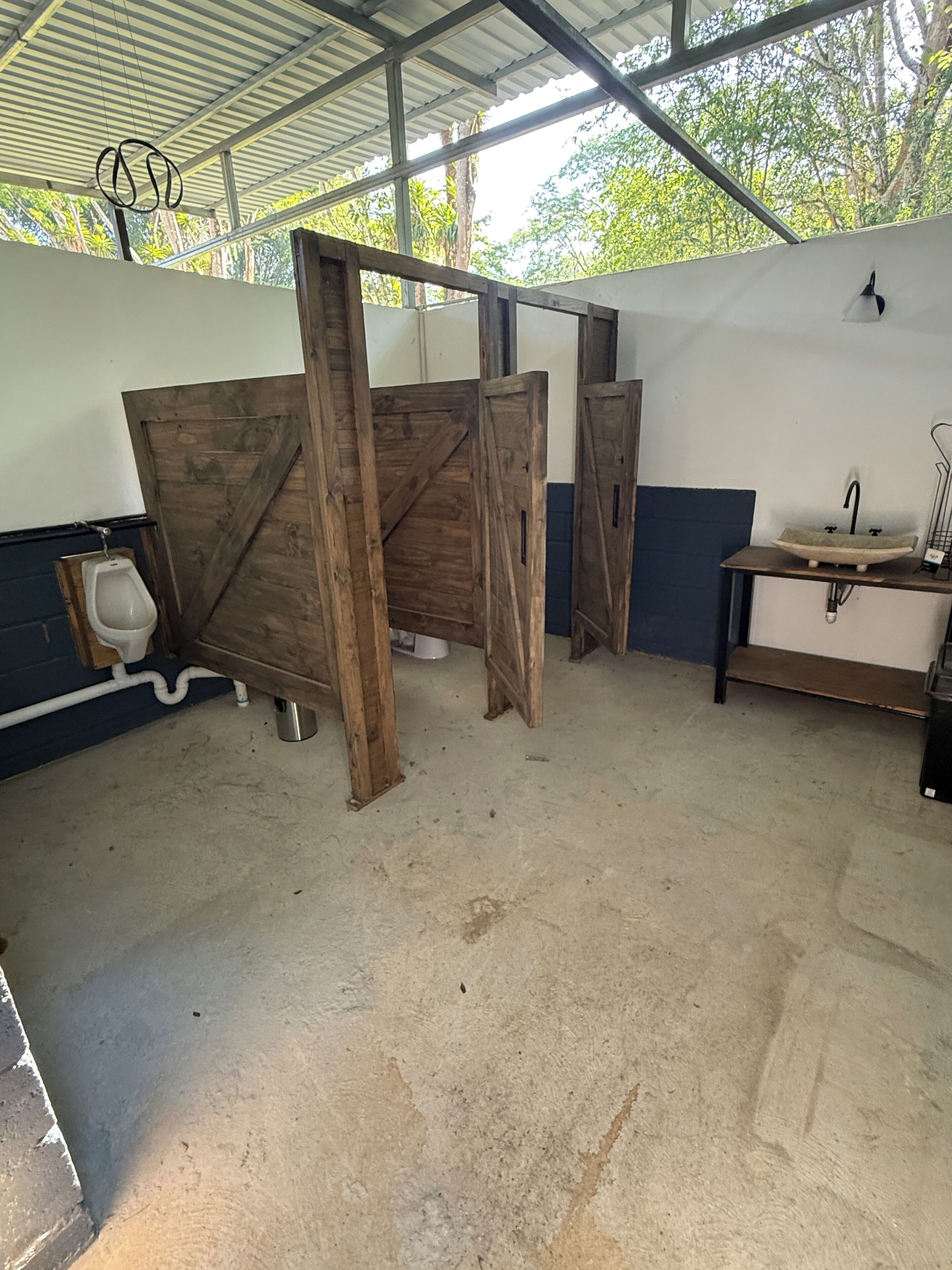 A restroom with wooden partitions, a urinal, a sink with a basin, and a metal rack.