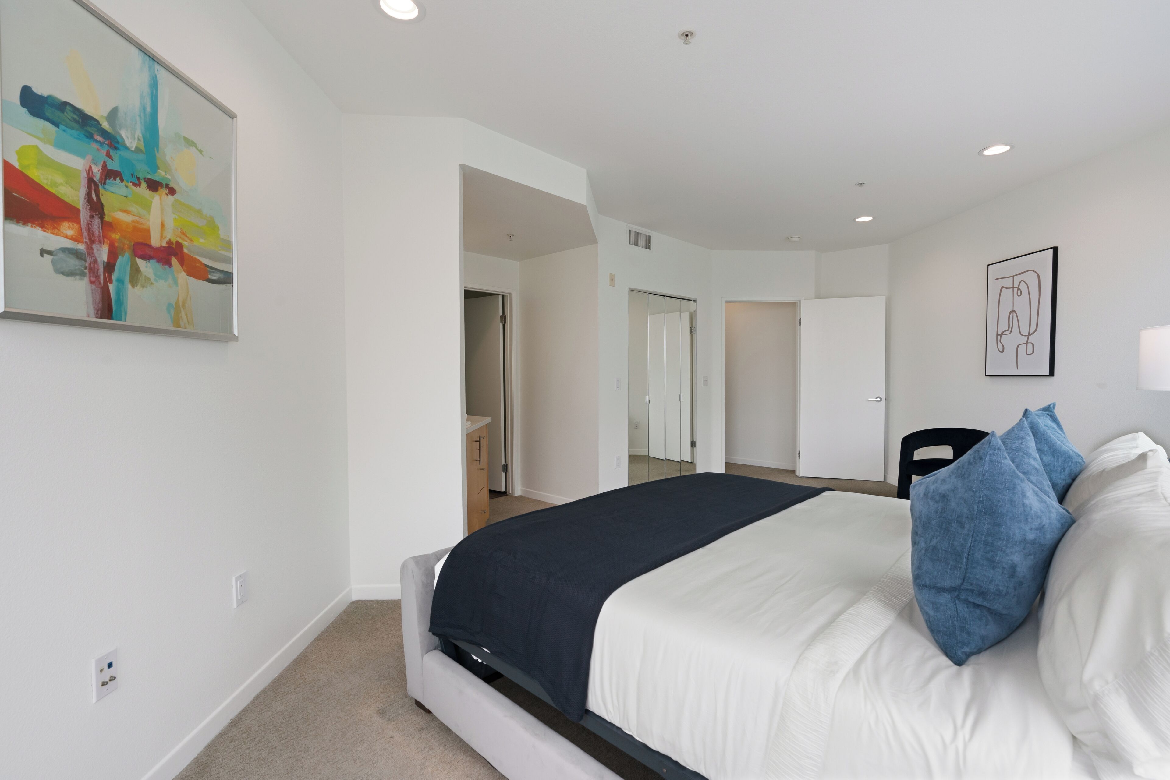 A modern bedroom with a bed, a painting, and a door leading to another room.