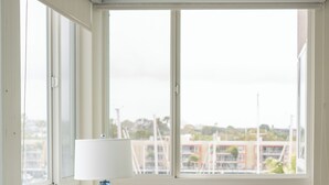 Une chambre d’hôtel avec une grande fenêtre, un lit avec une literie blanche, une lampe bleue et une vue sur un port de plaisance.