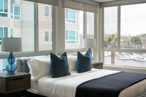 Une chambre d’hôtel avec un lit, deux tables de chevet, deux lampes et une vue sur une marina, visible à travers de grandes fenêtres.