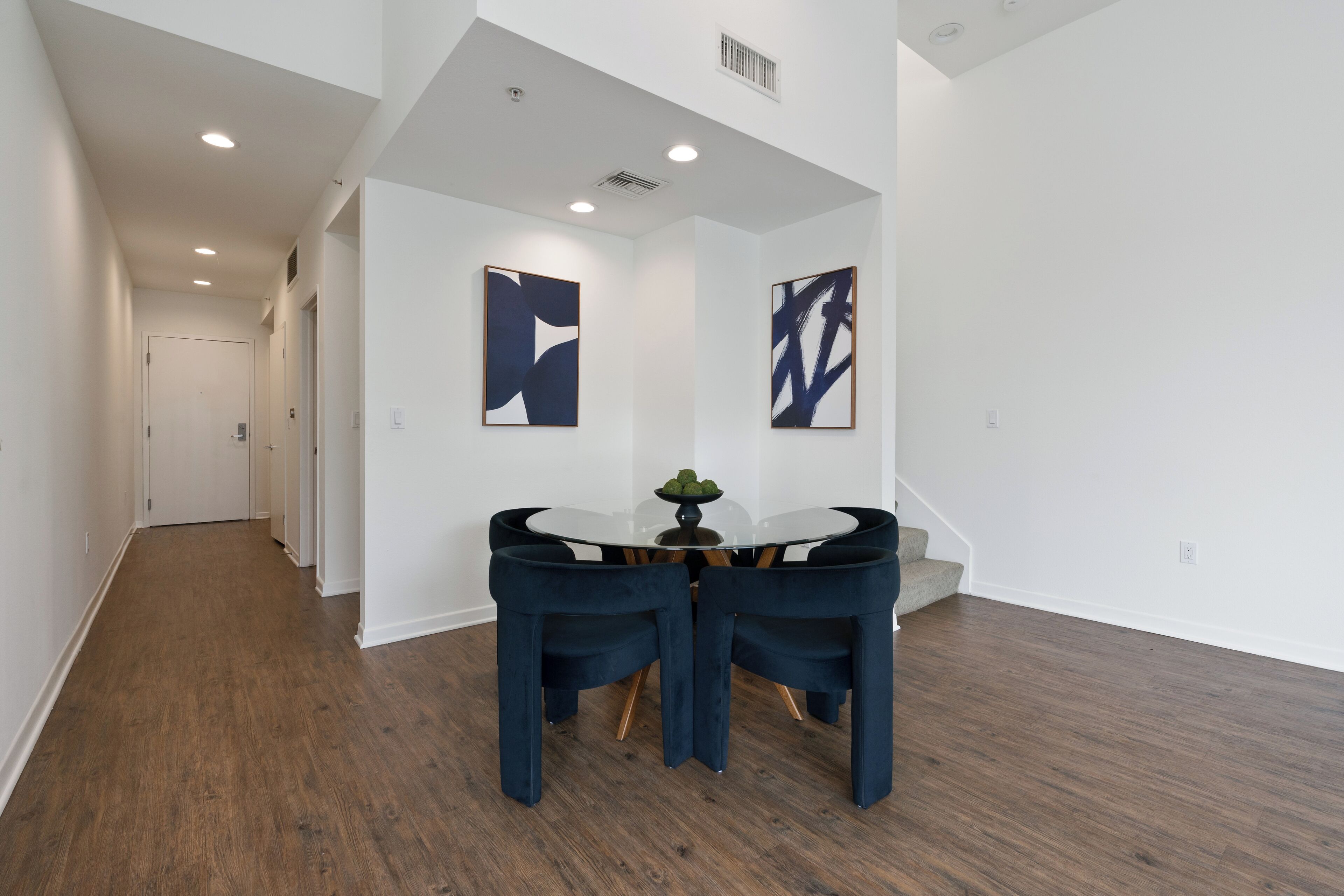 Un intérieur moderne avec un coin repas comprenant une table ronde et des chaises foncées, un couloir avec une porte, et deux tableaux abstraits accrochés aux murs.