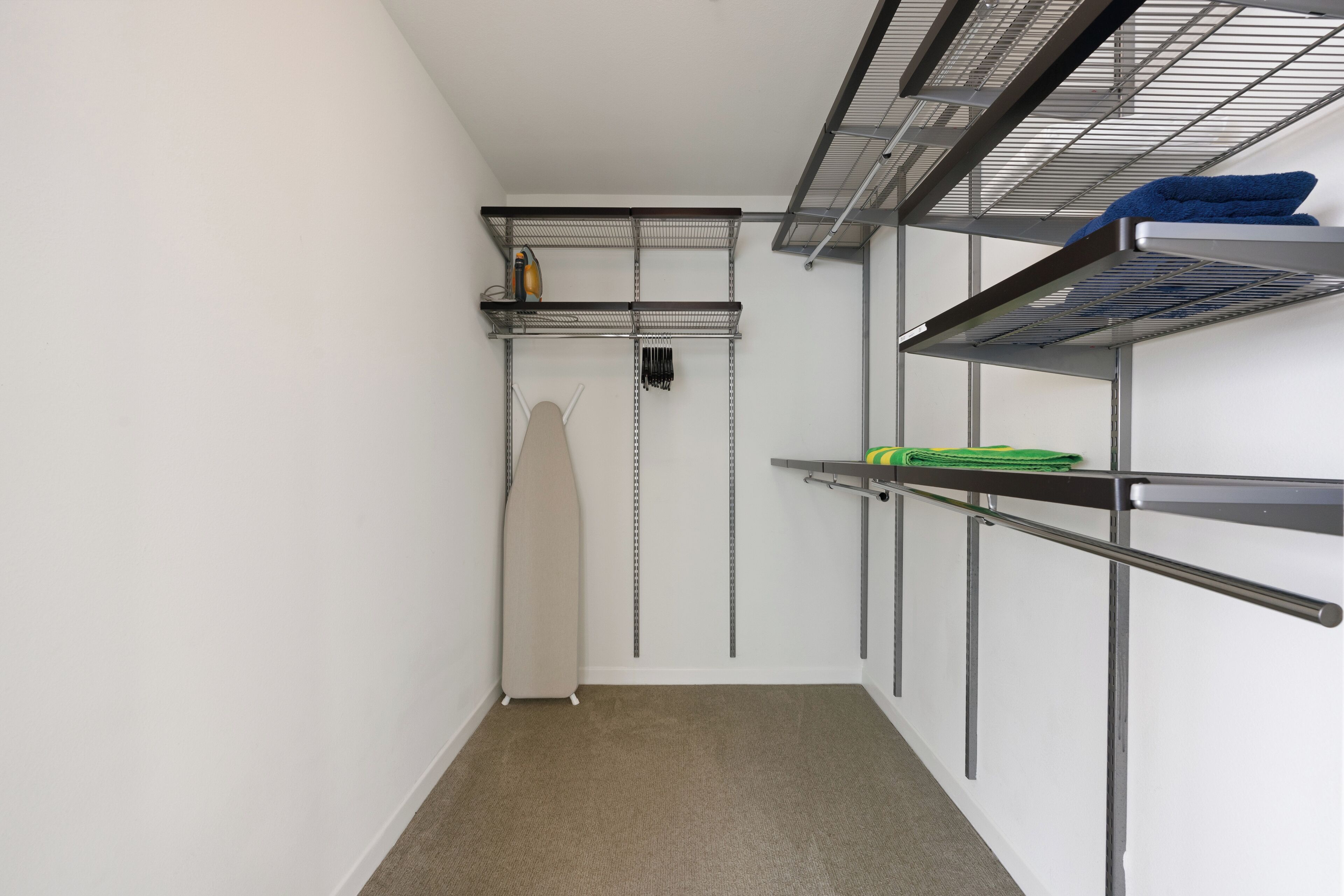 Un corridoio stretto con scaffali metallici, un asse da stiro color beige e una porta con maniglia nera.