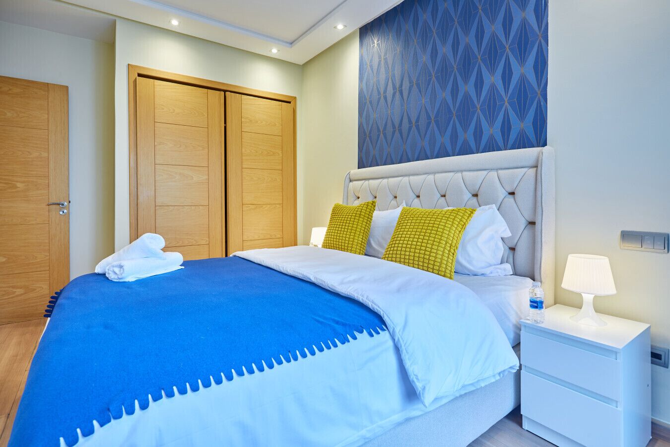 Quarto de hotel com cama, mesa de cabeceira, abajur e parede com padrão geométrico.