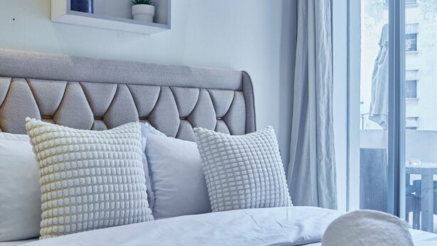 A neatly made bed with pillows, a bedside shelf with a plant, and a window with curtains.