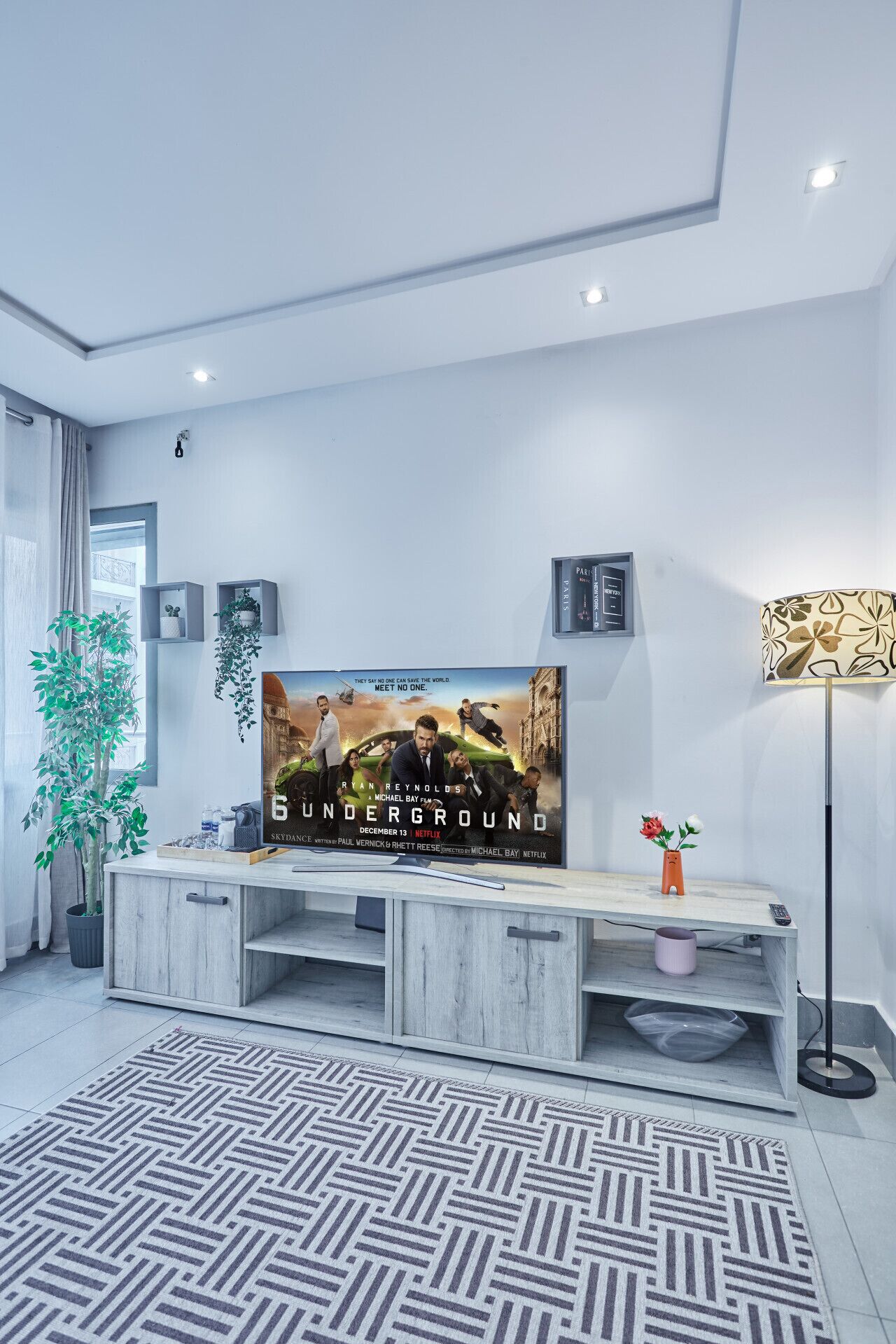 Un salon moderne doté d’un grand téléviseur à écran plat, d’un meuble de rangement en bois, d’une plante en pot, d’un lampadaire et d’un tapis à motifs.