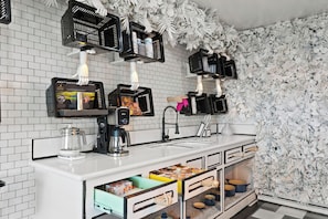 A modern kitchen with white tiled backsplash, black hanging baskets, and a decorative leaf wall. - LA Comic Mayhem (Long Beach)