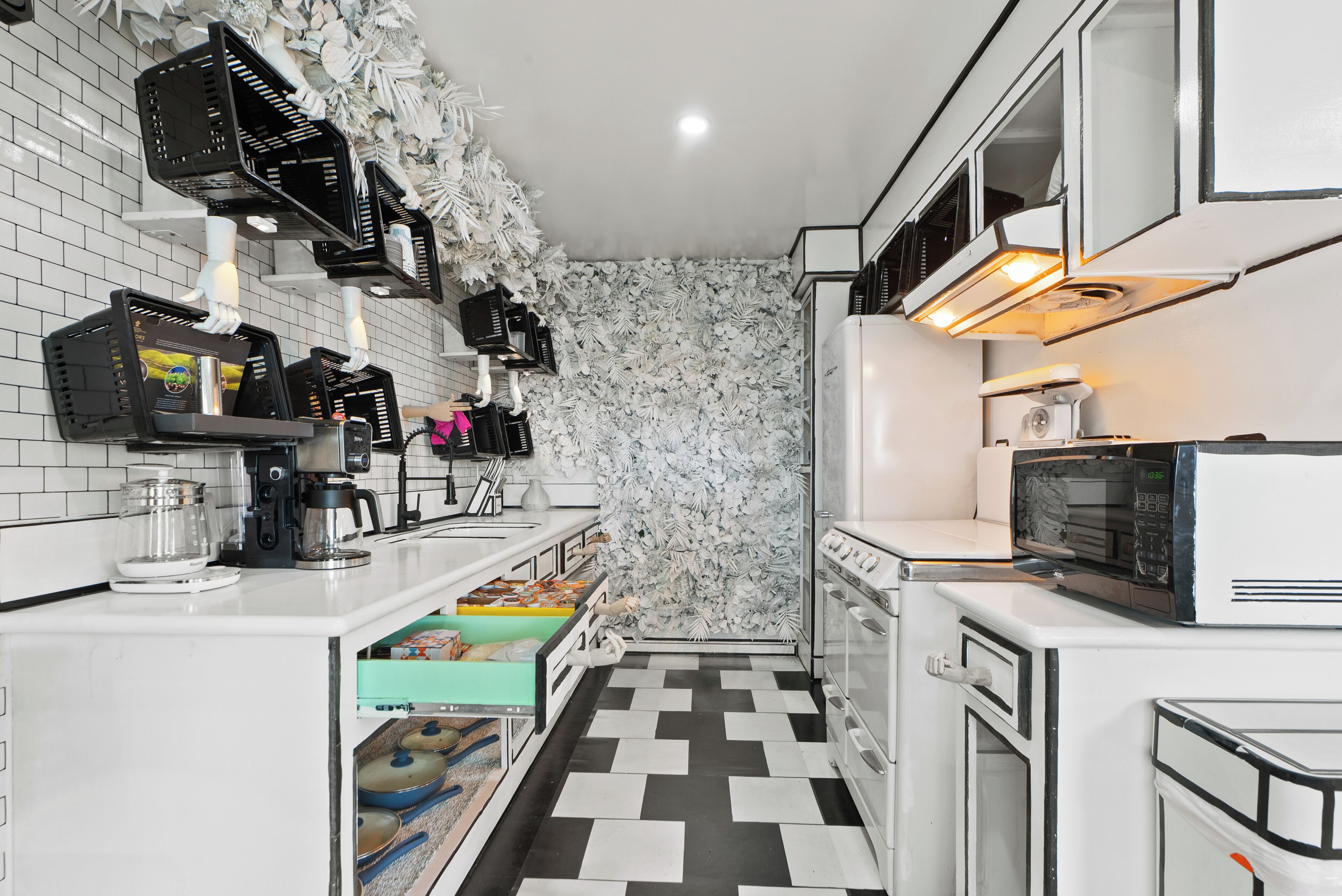 Une cuisine de taille réduite avec des meubles blancs, un carrelage noir et blanc et un mur décoratif orné d’objets suspendus.