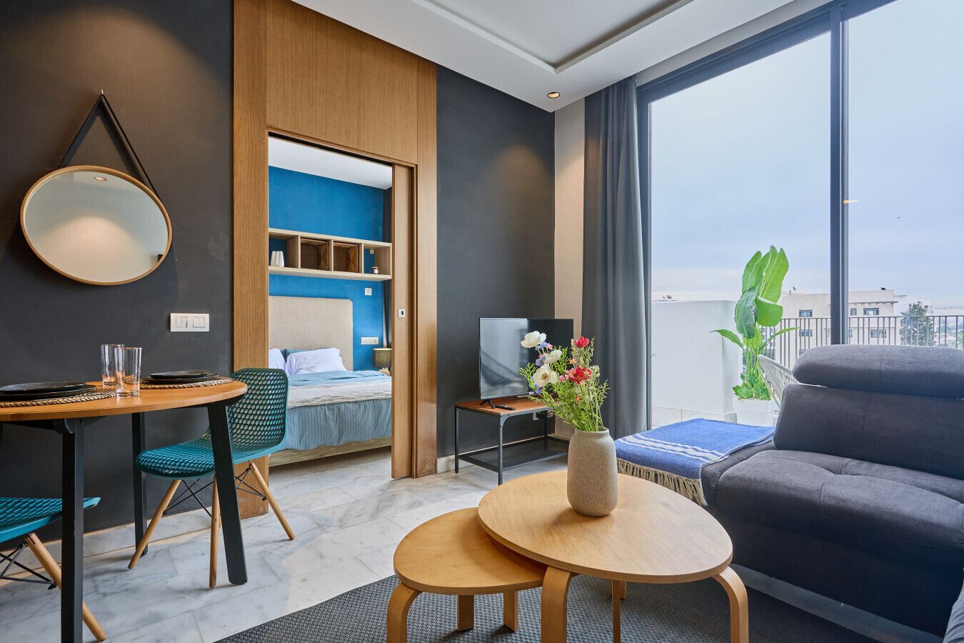 A modern hotel room with a bed, a dining area, a TV, a small table with a vase of flowers, and a large window with a view.