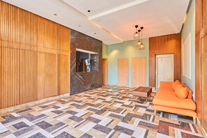 Quarto de hotel com piso em padrão herringbone, paredes revestidas de madeira, sofá com almofadas laranja e luminária pendente.