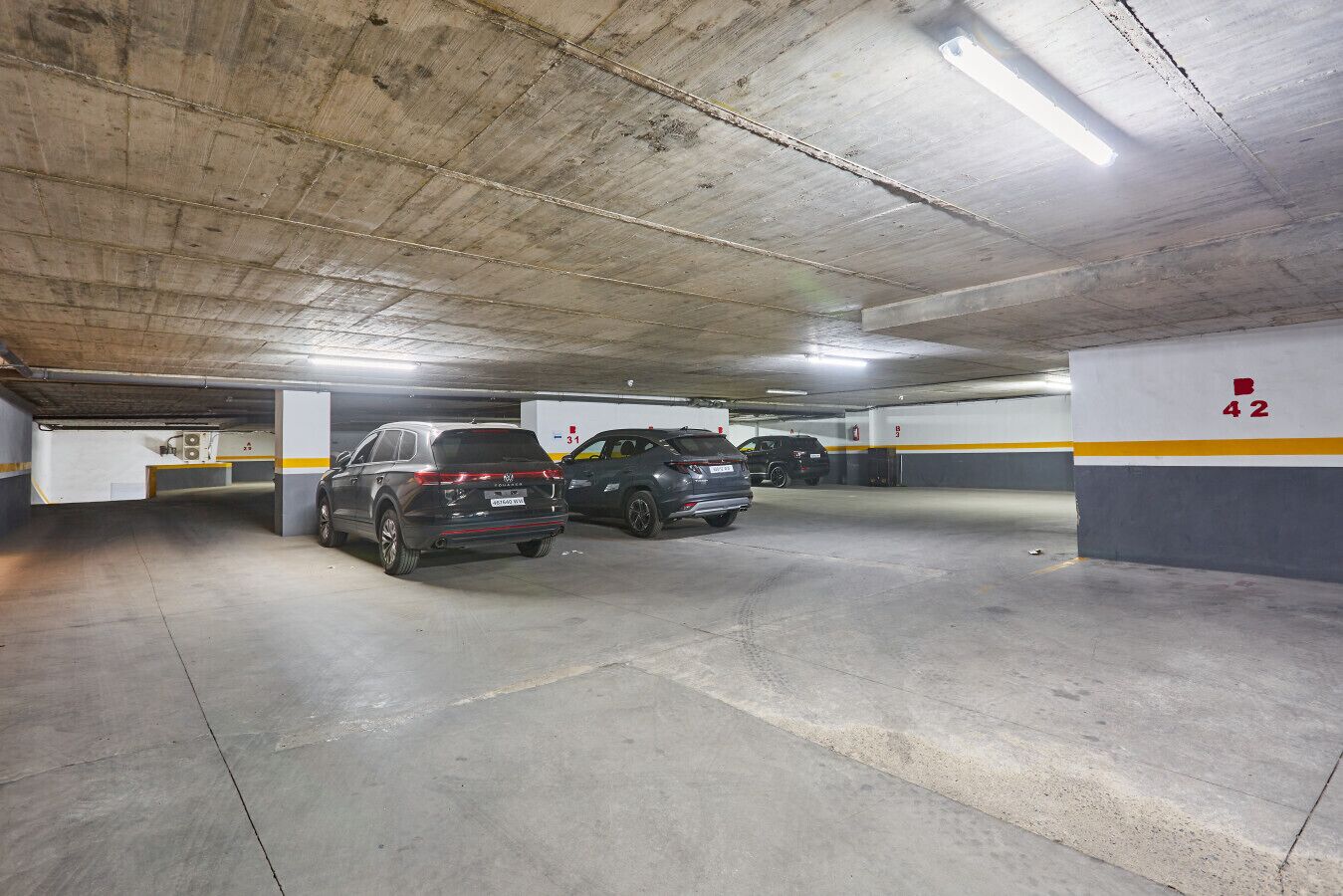 En parkeringskælder med flere niveauer, beton gulve og vægge, afmærkede parkeringspladser og belysning over hovedet.