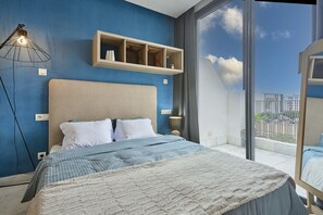 Quarto com uma cama grande, uma mesinha de cabeceira, prateleira na parede, luminária de chão e varanda com vista.