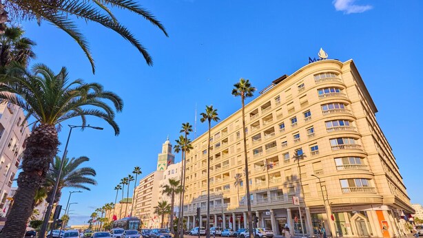 Un hotel de varios pisos con un minarete destacado, palmeras y un pavimento a cuadros.
