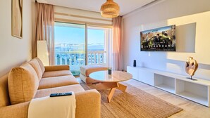 Una sala de estar moderna con un sofá, una mesa redonda de centro, un televisor montado en la pared y un ventanal con vistas.