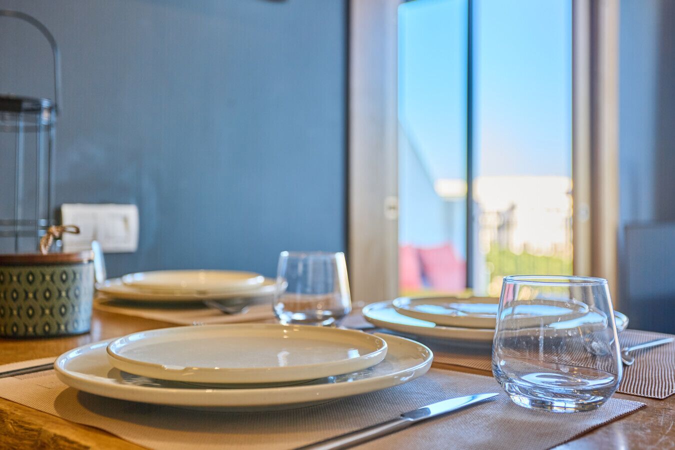 A dining table set with plates, glasses, and a pitcher, with a window in the background.