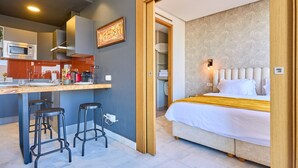Une chambre d’hôtel moderne équipée d’un lit, d’une kitchenette et d’une salle de bain, le tout visible à travers une porte ouverte.