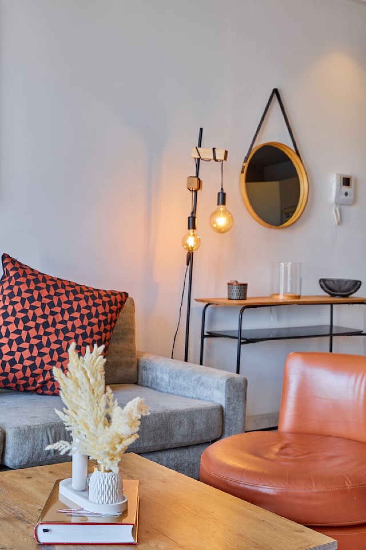 Un salon moderne comprenant un canapé, une table basse avec un livre et un vase, un fauteuil en cuir, un miroir et une lampe sur pied.