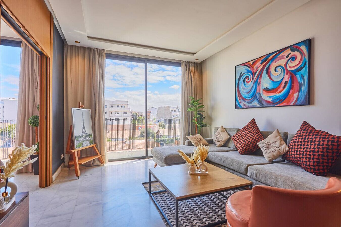 A modern living room with a large window, a painting of the Eiffel Tower, a sofa with patterned cushions, and a colorful abstract painting on the wall.