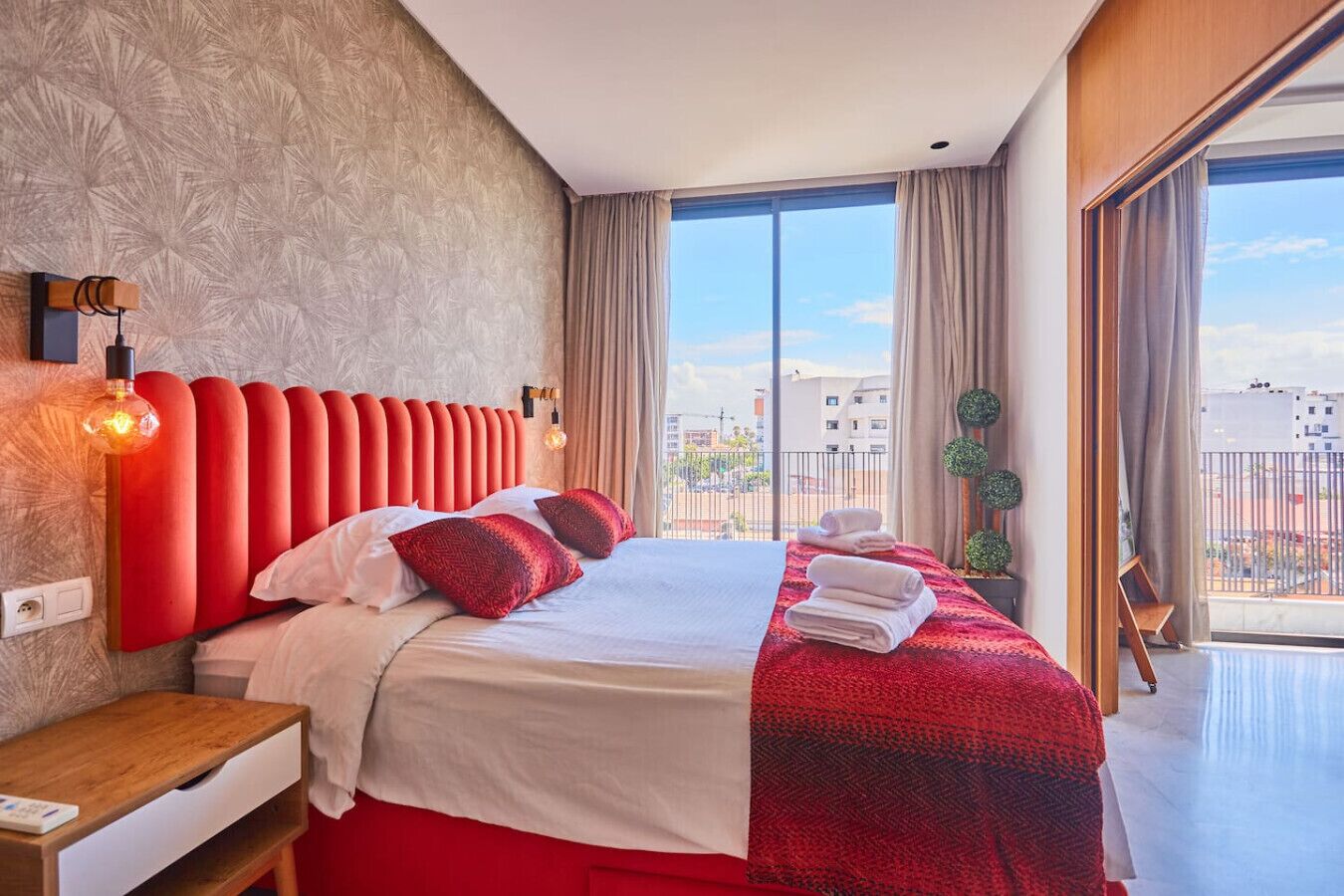 Una habitación de hotel con una cama grande, una mesita de noche, un cabecero rojo, una ventana con vistas a edificios y un balcón.