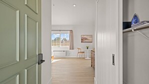 En hall med en grön dörr, ett vitt element, ett fönster med gardiner, ett litet bord med en stol och en tavla på väggen.