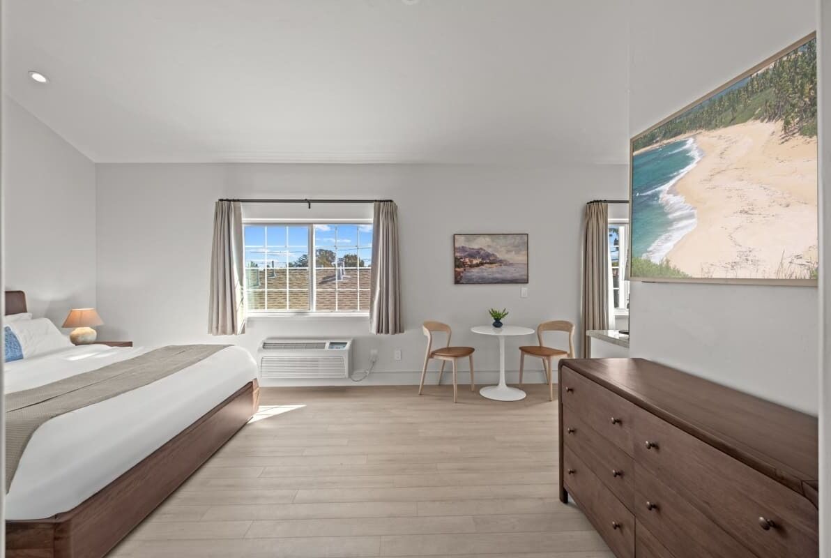 Habitación de hotel con cama, escritorio, silla, cómoda, ventana con cortinas y una pintura con vista al mar.