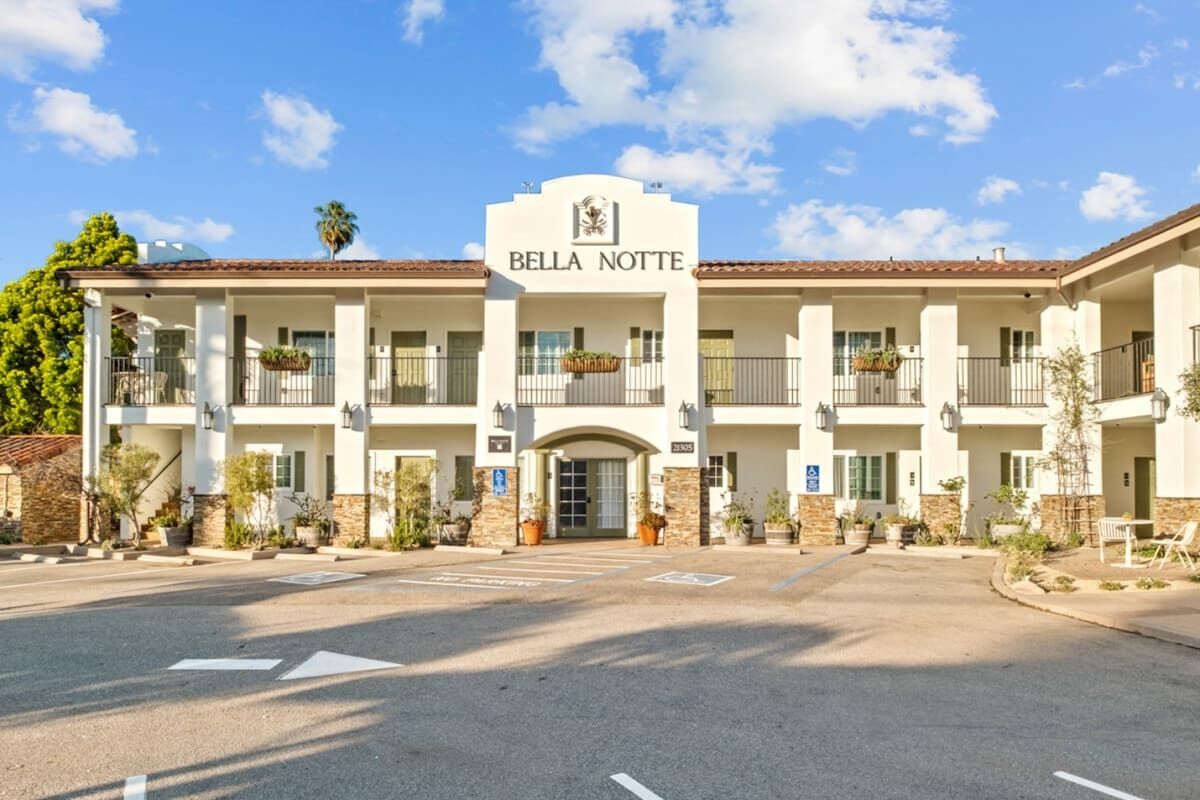 Een meerlaags hotel met een centrale ingang, balkons met beplanting en een heldere lucht.