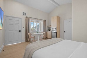 A bedroom with a bed, a small table, two chairs, a window with curtains, a kitchenette, and a door. - Bella Notte by the beach Studio 7 (Pleasure Point)