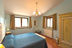 Ein Schlafzimmer mit einem Bett, zwei Fenstern mit Vorhängen, einem Heizkörper, einem Spiegel, einer Kommode und einer Lampe.