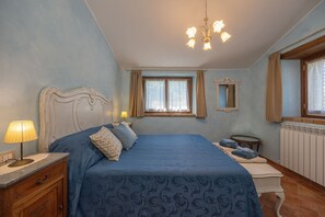 A bedroom with a large bed, a bedside table with a lamp, a window with curtains, a mirror, and a radiator. - Agriturismo Antica Quercia Verde - Gonfianuvole (Cortona)