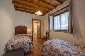Une chambre avec deux lits, des poutres apparentes en bois au plafond, une fenêtre avec des rideaux et une porte donnant sur une autre pièce.